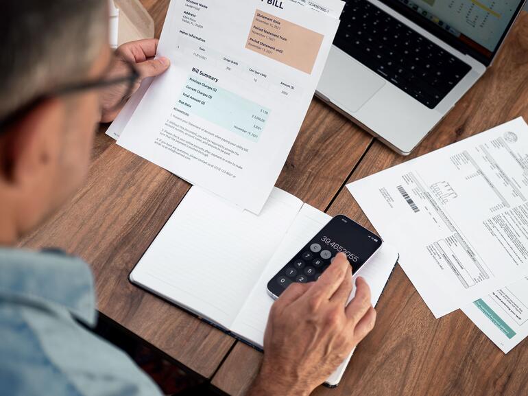 Man calculating utility expenses using a smartphone at a wooden desk.