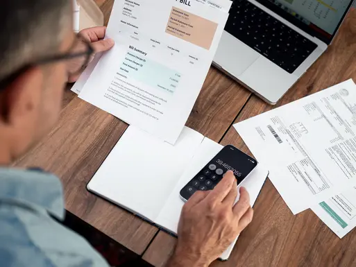 Man calculating utility expenses using a smartphone at a wooden desk.